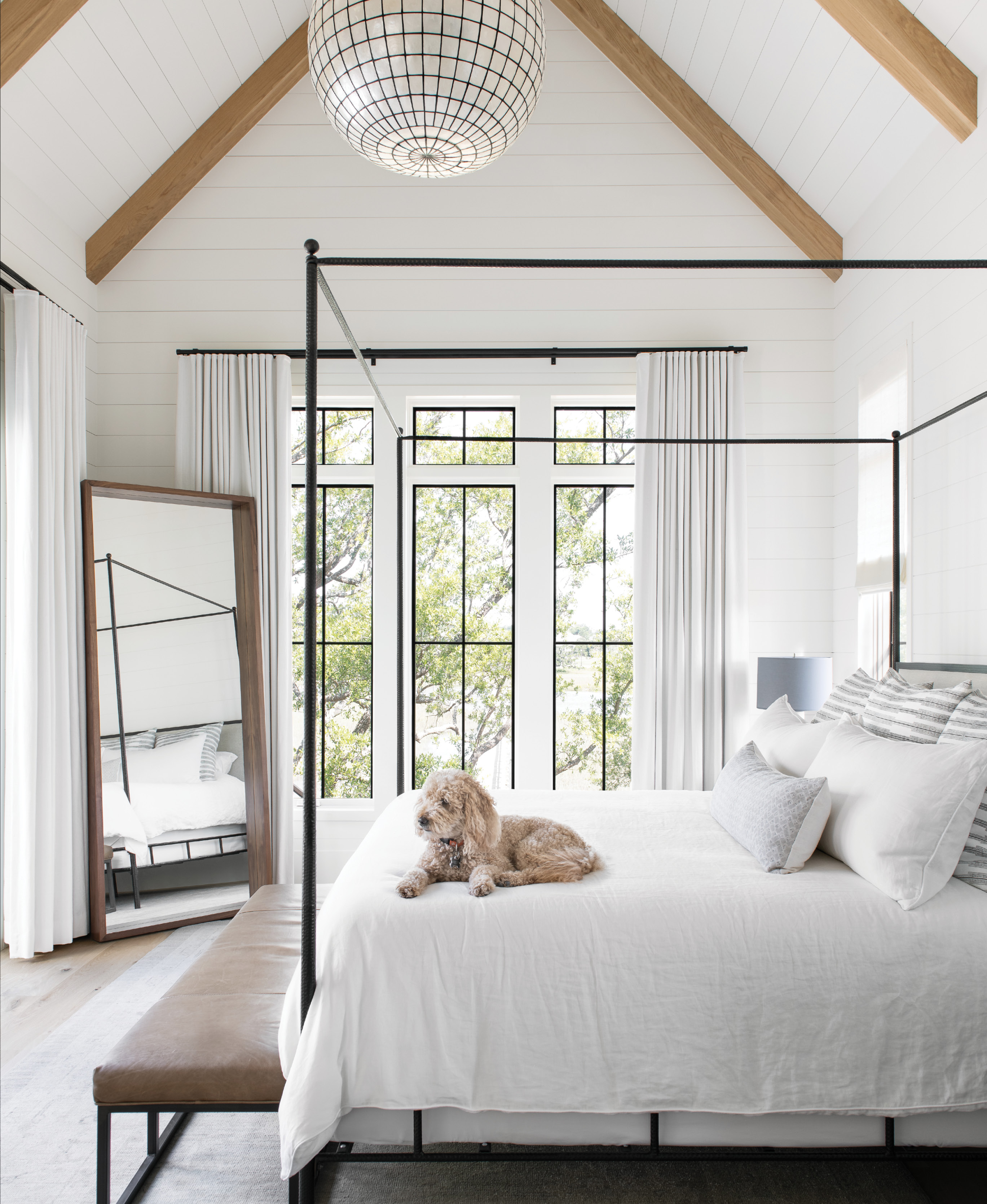 Simply Luxe: The paired-down master suite feels glamorous yet comfortable. Tying into the black steel of the windows, the iron-framed “Marco Bed” by Oly Studio is softened by plush Peacock Alley bedding and a worn leather bench atop a vintage Oushak rug. A Restoration Hardware chandelier and Room &amp; Board mirror complete the space.