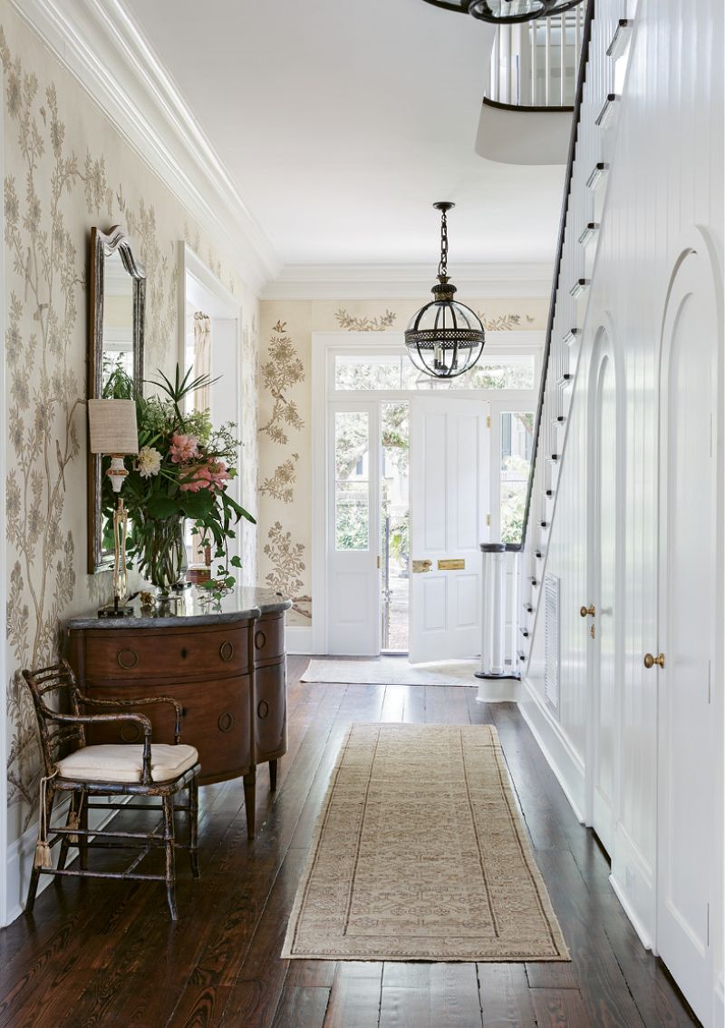 HAUTE ARRIVAL: The entrance hall adds a dash of drama courtesy of exquisite Gracie “Parchment Vista” wallpaper. Shades of cream together with gilt and silver produce a mesmerizing effect, complemented by the curves and scale of a circa-1850 stone-topped Italian buffet from Foxglove Antiques. Two English Regency armchairs from Charleston’s Golden &amp; Associates Antiques round out an eclectic mix of European pieces.