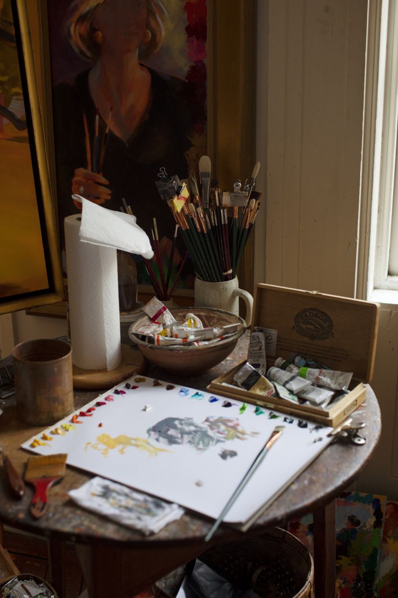 A ceramic mug filled with brushes and tubes of oil paint rest on a cricket table, layered in a paint-flecked patina after decades of use.
