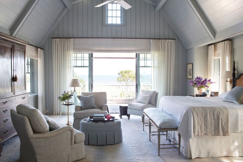 Sleeping bEauty: In the primary bedroom, decorative woodwork painted in Farrow &amp; Ball’s “Light Blue 22” evokes the deep blue ocean beyond the picture frame window. Sheer drapes allow the sunlight to stream through and comfy club chairs from TCS Designs pair with an antique armoire and bed for a touch of relaxed luxury. An ottoman wrapped in Rogers &amp; Goffigon fabric and a bench from Suzanne Rheinstein’s Hollyhock complete the look.