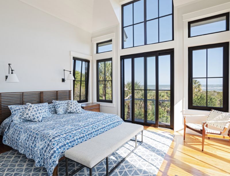 Soak it In: The primary bedroom is a simple space, where a wall of unshaded Henselstone tilt-and-turn windows, manufactured in Germany, allows the beauty of the surroundings to decorate the room and wake the couple naturally. “We’ve been getting in tune with nature out here,” says Toni. “We can tell what time it is by the position of the moon.”